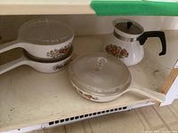 Photo of two white CorningWare sauce pots with long handles and clear glass lids, decorated with floral fruit patterns. Also includes matching tea pot with screw-in black handle and silver rim on lid.