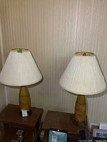 Pair of vintage table lamps shown side by side on wooden tables against striped wallpaper, both with textured tan ceramic bases and white pleated shades.