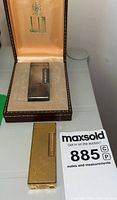 Silver-finish lighter in open Dunhill box alongside gold-finish lighter and auction card