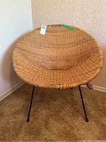 Front view of a rounded wicker chair with a deep bowl-shaped seat and three black metal legs on a beige carpeted floor.