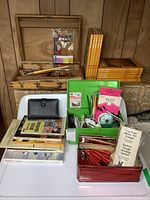 Collection of wood carving tools in wooden box, craft books stacked, green artist kit open with tools, and other craft items arranged on a white chair and in front of a wooden cabinet.