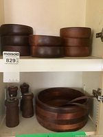 Eight small wooden salad bowls and two sets of wooden salt and pepper grinders on a white shelf, large wooden salad bowl with serving utensils below.