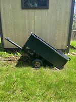 Side profile of black metal yard trailer with 33x44 inch bed, tilted bed, flat tires, and metal frame visible against grass and shed background.