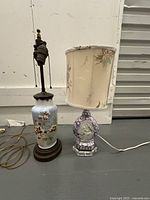 Pair of vintage table lamps showing one glass floral lamp with pull chains and no shade, and one ceramic cameo lamp with shade, both plugged in front of a gray wall.