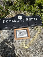 Front view of the black wooden Hotel de Paris wall rack with white lettering, four metal hooks, and a central clock, along with the small framed New Orleans street scene print, both displayed outdoors.