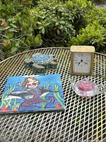 Photo of all four decorative items on outdoor metal mesh table: Tiffany & Co. brass desk clock, turtle trinket box with blue enamel, hand-painted mermaid ceramic tile, and miniature glass teapot with pink flower.