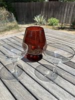 Photo of the deep amber brandy snifter alongside two clear glass coupes with silver-painted rims, displayed on an outdoor wooden surface.