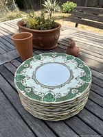 Stack of 10 vintage Noritake M dinner plates with emerald green and gold scroll borders viewed outdoors on wooden table.