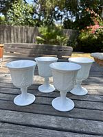 Four milk glass goblets with grapevine and leaf embossed pattern displayed on outdoor wooden table, showing overall lot composition and detail.