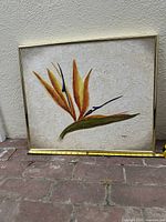 Front view of the framed textured Bird of Paradise flower painting showing vivid orange, gold, and purple colors on a cream textured background. Signature 'Cooper' is visible on lower right corner. A yellow measuring tape laid in front indicates approximate width.