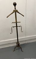 Front angled view of the vintage brass valet stand showing the hanger top, accessory tray, curved arms, and tripod base.