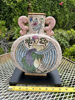 Front view of vintage ceramic Chinese moon flask vase on a black wooden base showing raised phoenix design, floral and butterfly motifs, and sculptural pink handles. Measuring tape shows width approx 7.5 inches.