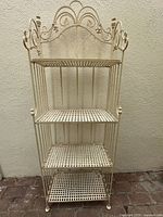 Full front view of the cream-colored metal baker's rack showing all four lattice shelves and decorative scrollwork and grape cluster detailing on the top.