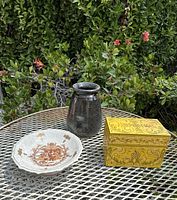 Photo showing all three vintage home décor items together on outdoor metal table with greenery background. Items include the footed crackle-glaze ceramic bowl with Rococo scene, yellow floral tin box with hinged lid, and dark speckled studio pottery vase.