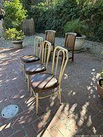 Three vintage metal frame cafe chairs arranged on brick patio by plants, showing cream tubular arched backs and brown vinyl seats in sunlight.