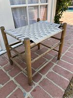Side view of vintage wooden Shaker stool showing round legs, dowel stretchers, and woven fabric webbing seat in worn condition.