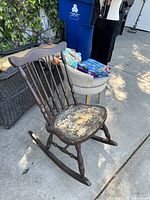 Vintage dark brown wooden rocking chair outdoors on pavement, showing spindle-back and floral upholstery on seat.