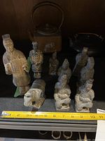 Eight small warrior statues in light gray ceramic material with one small ceramic elephant and two tea pots visible behind them, with measuring tape showing about 5 inches tall for tallest statue.