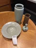 Four ceramic items displayed: three vases including tall pale blue design with embossed floral patterns, small pale blue bottle-shaped vase with pink flower, medium brown glazed vase; plus large shallow serving bowl with speckled tan glaze, all on wooden surface.