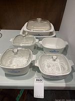 Full set of six white CorningWare dishes with blue floral decoration displayed with lids on three and others open. Handles are visible on dishes.