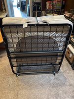 Rear view of folded bed frame with mattress secured by strap, showing metal wire grid and wheels.