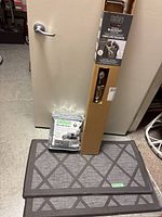 Shot of curtain rod box standing upright with packaging showing the nickel adjustable curtain rod information, two memory foam mats with geometric design on the floor, and a packaged grey sofa cover leaning against a door.