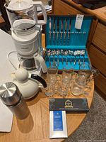 Overall shot showing all items including cutlery set in turquoise case, coffee maker with carafe, white thermos, glass teapot, glasses, and tea box on table.