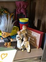 Image showing three Willow Tree-style figurines, a troll doll with gray hair, a decorative box with motivational quote, and colorful plastic containers in background.