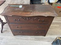 Solid wood dresser front view showing three drawers with ornate metal handles and wood carving detail, surface wear visible.