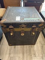 Front view of vintage black hat box chest showing two side locks and broken central lock, metal rivets, and corner reinforcements.