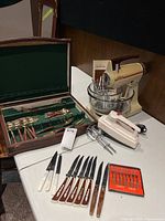 Full view of the lot showing the Sunbeam Mixmaster stand mixer with glass mixing bowls, Sunbeam handheld mixer, wooden box cutlery set, knives, and small red boxed set.