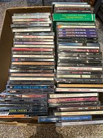 Wide shot showing a large box filled with stacked used music CDs and some cassettes, including visible titles such as Santana, Dixie Chicks, Barbra Streisand, Leonard Cohen, and more.