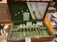 Opened wood chest showing silver plated cutlery set arranged in slots on green velvet lining. Two serving utensils mounted in lid. Chest top interior has labels for 1881 Rogers pattern and certificate.