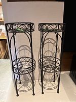 Front view of two black metal plant stands with round open wire shelves and decorative scrollwork on top edge.