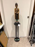 Full view of the wooden Buddha statue on the black metal stand against a wall.