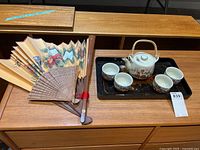 Wide view of Asian inspired tea set on black serving tray, with two decorative fans placed beside it on wooden table.
