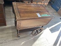 Photo showing full view of the solid wood tea trolley with carved detailing and large ornate wooden wheels on a light wood floor.
