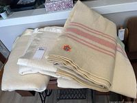 Three folded blankets on a wooden bench: pink-striped blanket atop two plain off-white blankets