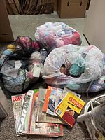 Three mesh bags filled with assorted yarn skeins in multiple colors, accompanied by a small stack of knitting and crochet books spread in front.