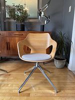 Front view of the Olé wooden-shell chair showing the curved shell, cut-out armrests, and light gray upholstered cushion on polished aluminum swivel base.