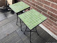 Two vintage side tables with green mosaic tile tops and black iron bases, placed side by side on a textured floor next to a brick wall.