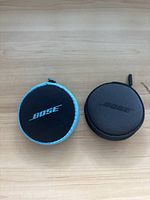Two small round Bose branded zippered cases placed closed on a wooden surface, one with black and blue trim, the other fully black.
