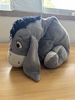 Side view of the gray donkey plushie, showing its black mane, large floppy ears, and embroidered facial features. Positioned sitting on wooden floor near a window.