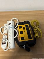 Photo of three extension cords and power strips on a wooden surface, showing white power strip with six outlets, yellow/black surge protector, and small white extension cord
