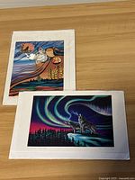 Two colorful Indigenous art prints on a wooden surface, one showing a wolf howling under aurora borealis, the other with stylized native animal and abstract designs.