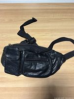 Black leather fanny pack with several compartments and adjustable strap displayed on light wood surface.