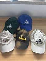 Five baseball caps in various colors and conditions on a tabletop — green, blue, white/beige, and distressed black with yellow branding.