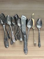 Full view of the old silverware laid out on a wooden surface showing different types and sizes of spoons, a butter knife, and a serving spatula.