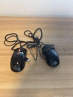 Two black wired SteelSeries computer mice, placed on a wooden surface. Both have USB cables, one with orange side buttons visible.