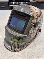 Front and side view of the welding helmet showing skull artwork, rectangular auto-darkening lens, and adjustment knob.